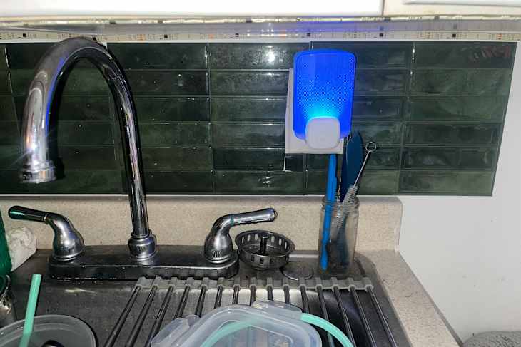 Kitchen sink area with a chrome faucet, blue light fixture, and utensils in a jar against green tile backsplash.