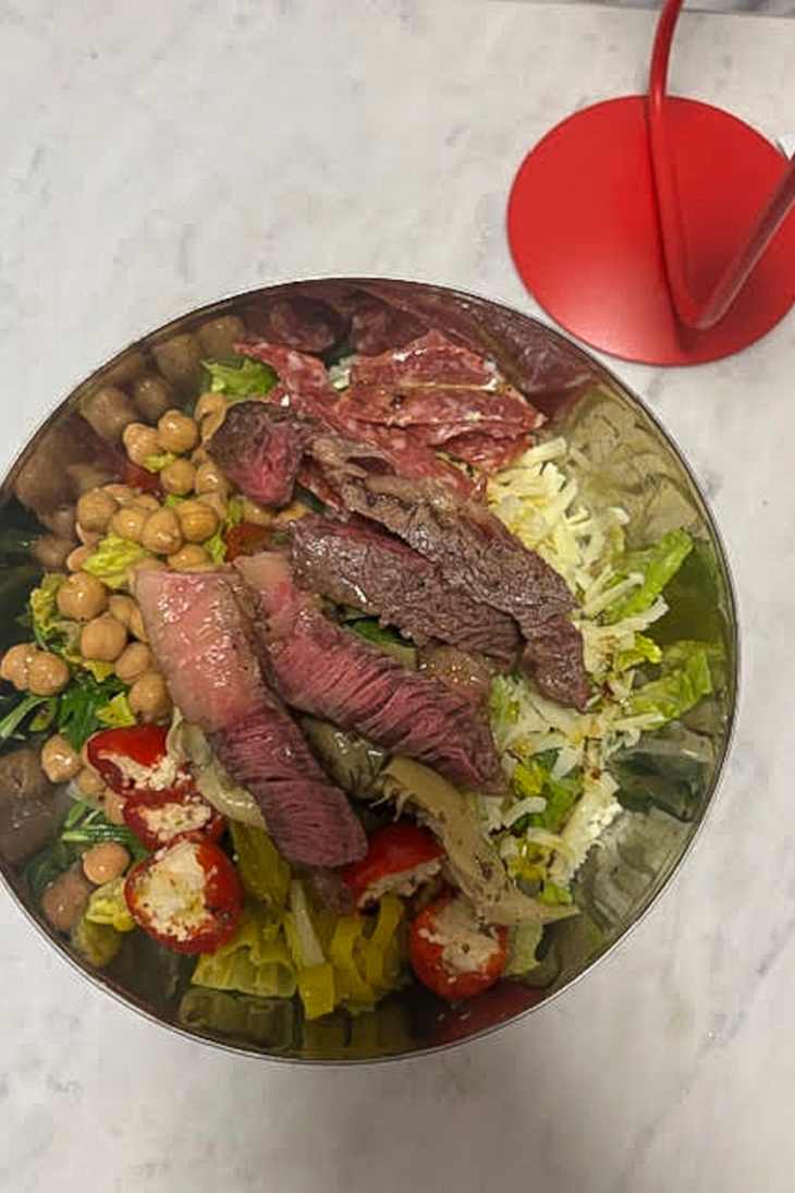 A large bowl filled with mixed greens, sliced steak, cherry tomatoes, chickpeas, and shredded cheese, with a red stand nearby.