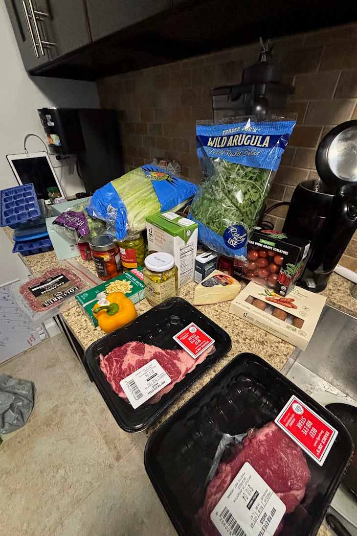 Assorted groceries on a kitchen counter, including arugula, celery, bell pepper, canned goods, and packaged meats.
