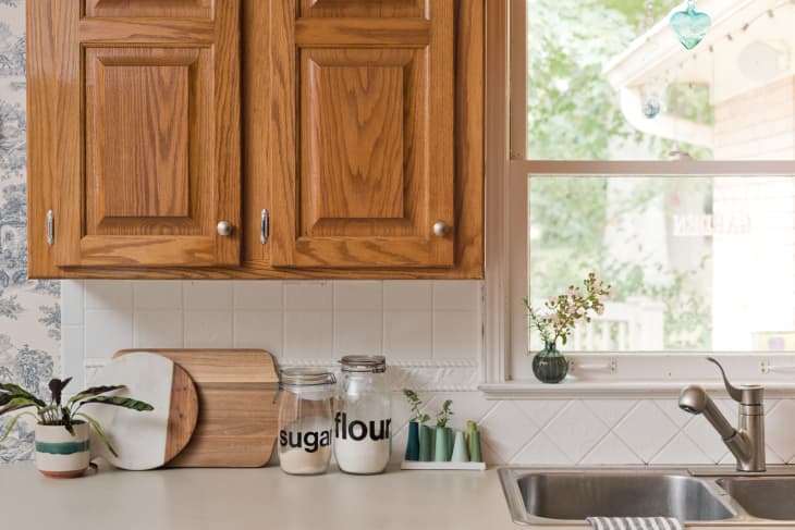 Kitchen counter with wooden cabinets, cutting boards, jars labeled sugar and flour, small plants, and a sink by a window.