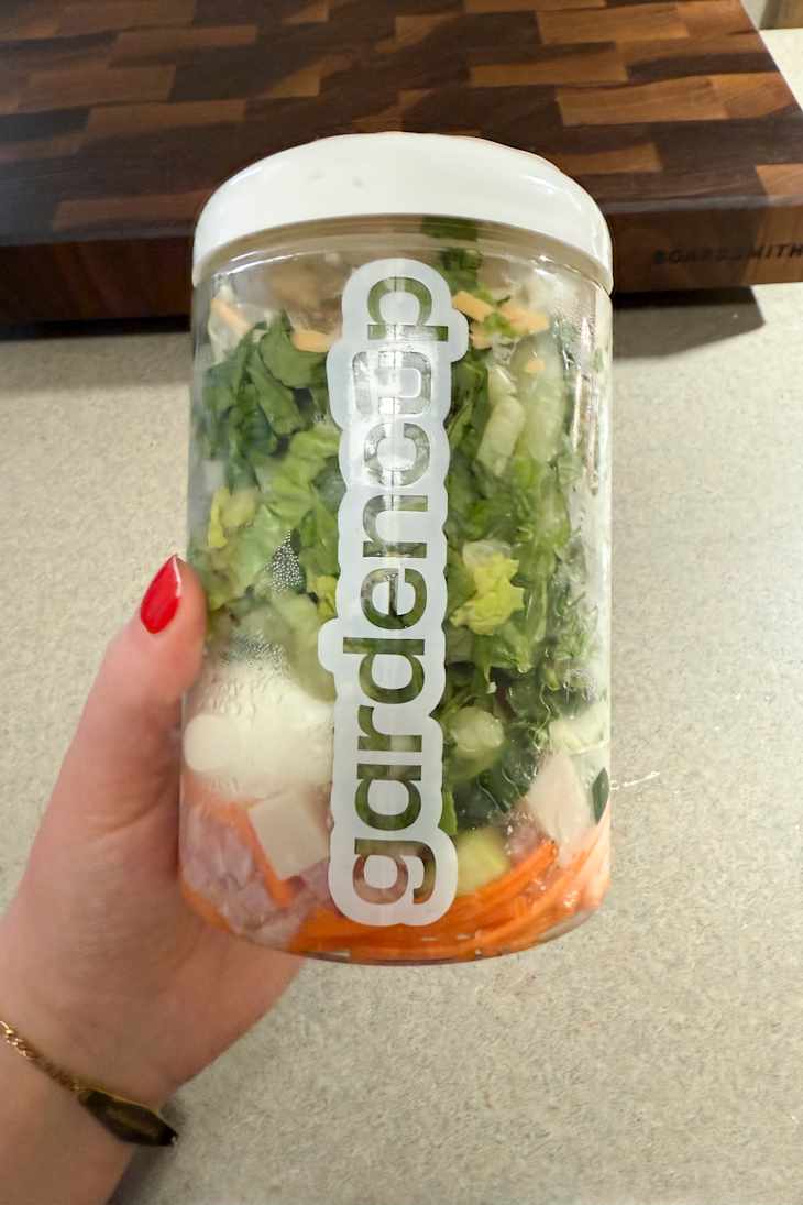 A hand holding a clear container filled with layered fresh vegetables, including lettuce, carrots, and cucumbers.