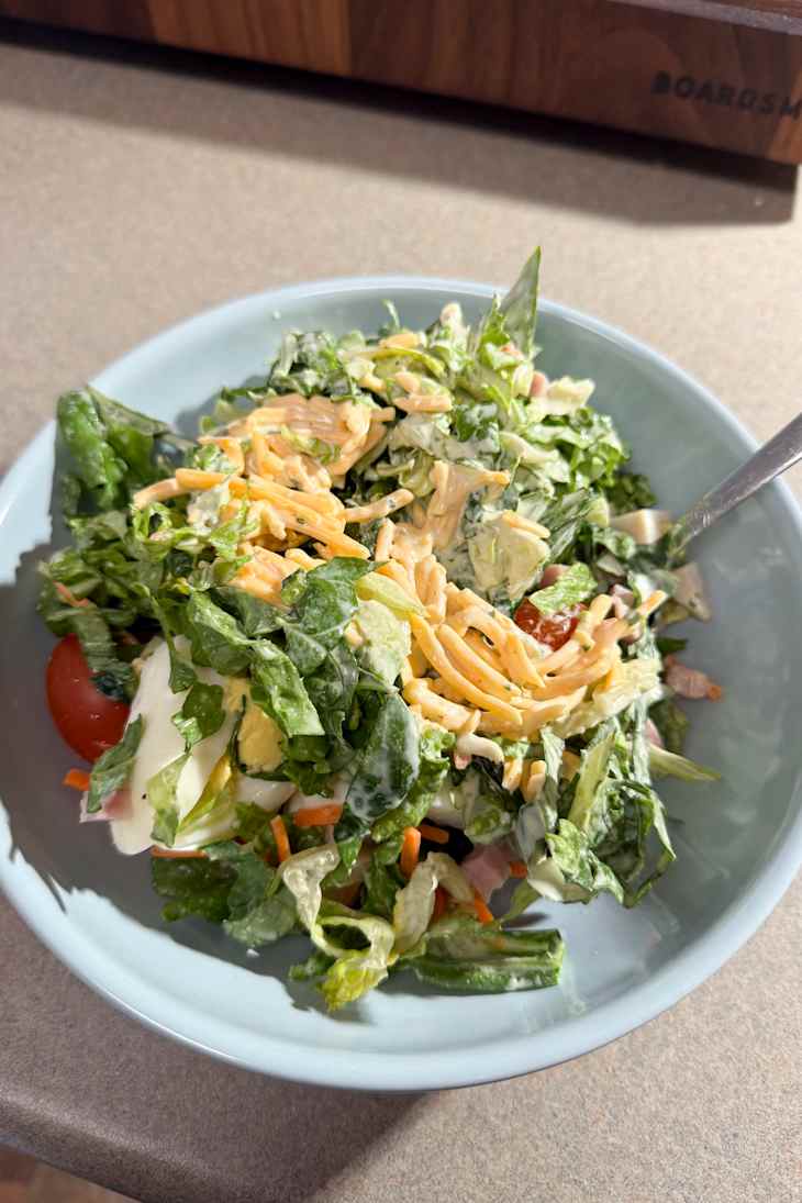 A bowl of mixed salad with greens, cherry tomatoes, shredded cheese, and creamy dressing.