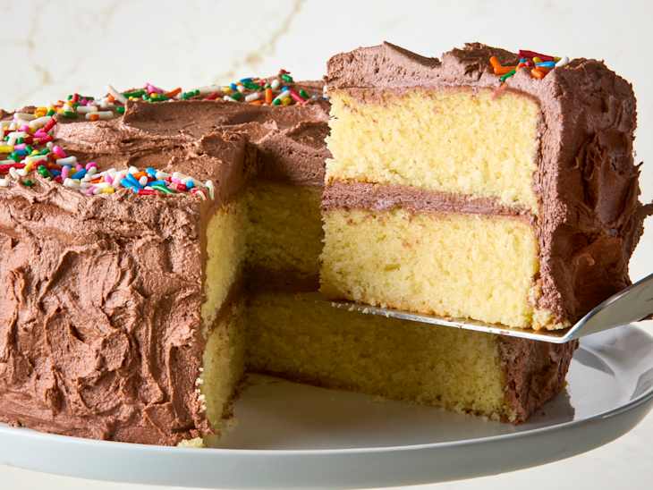 head on shot of a slice of yellow cake with chocolate icing being pulled from a full cake