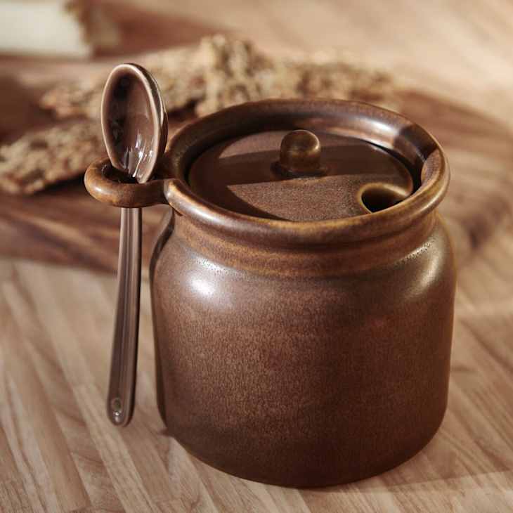 Brown ceramic jar with a lid and a spoon resting on the side, placed on a wooden surface.