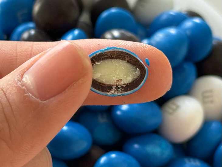 Someone holding a halved Cookies and Cream M&M over a bowl of candy