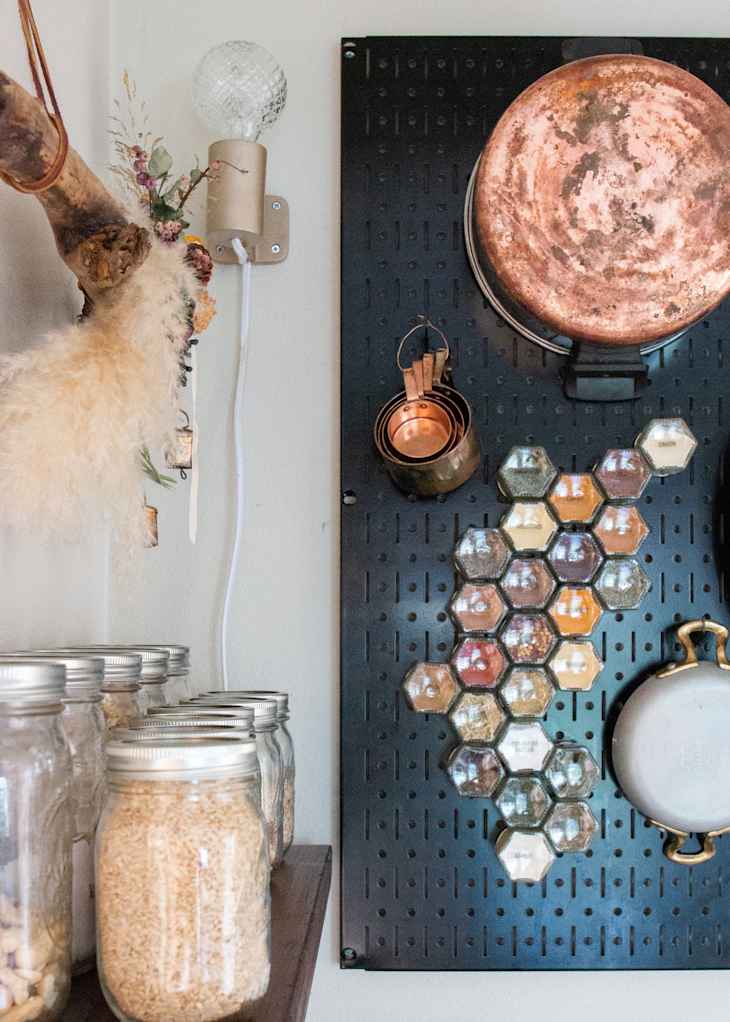 Jars of grains on a shelf beside a black pegboard with copper pots and hexagonal spice jars.
