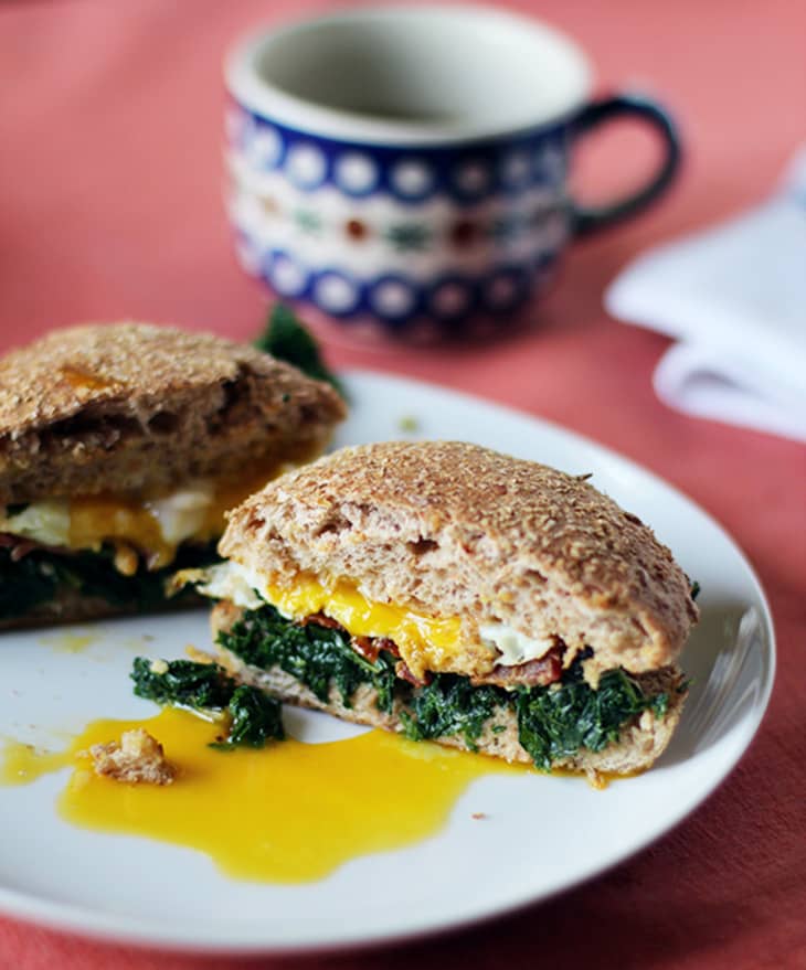 Whole grain breakfast sandwich with egg, spinach, and melted cheese on a white plate, next to a patterned mug.