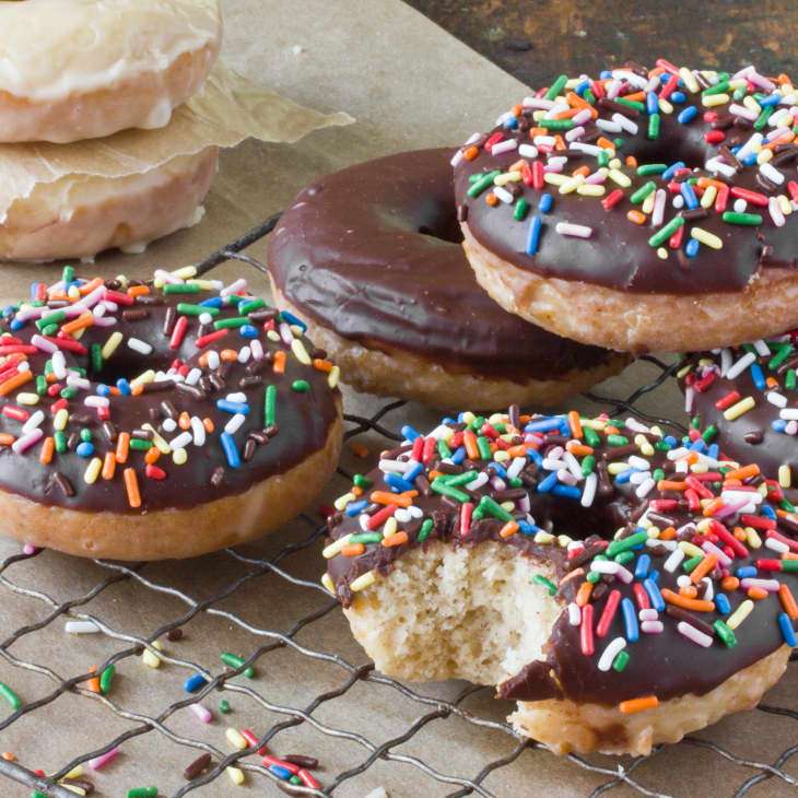 Chocolate glazed donuts with colorful sprinkles on a wire rack, one with a bite taken out.