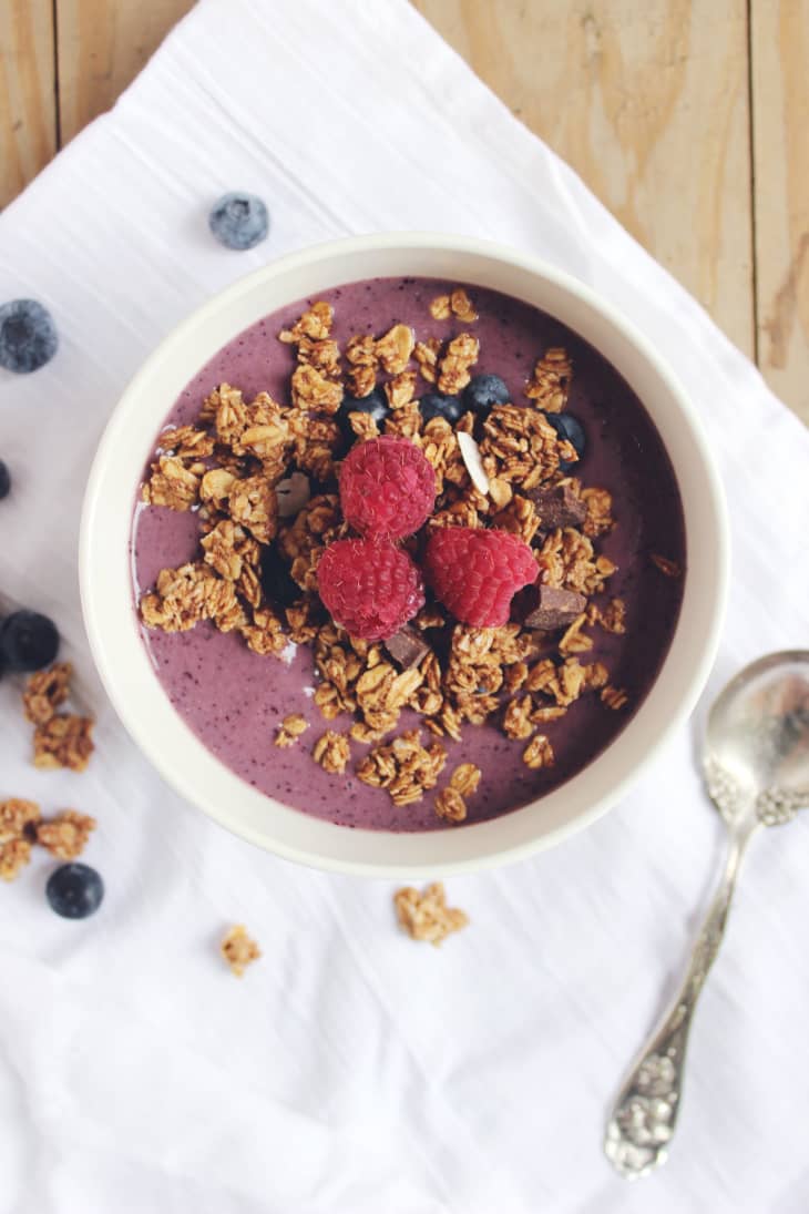 Berry smoothie bowl topped with granola, raspberries, and blueberries on a white cloth with a spoon.