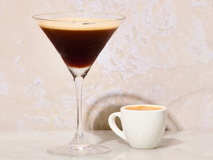A martini glass filled with dark coffee cocktail next to a small white espresso cup on a light marble surface.