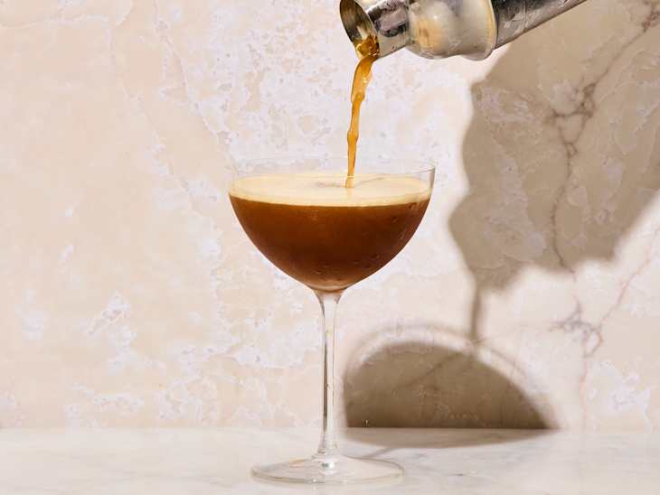 Cocktail being poured from a shaker into a coupe glass, with a creamy foam layer on top, against a marble background.
