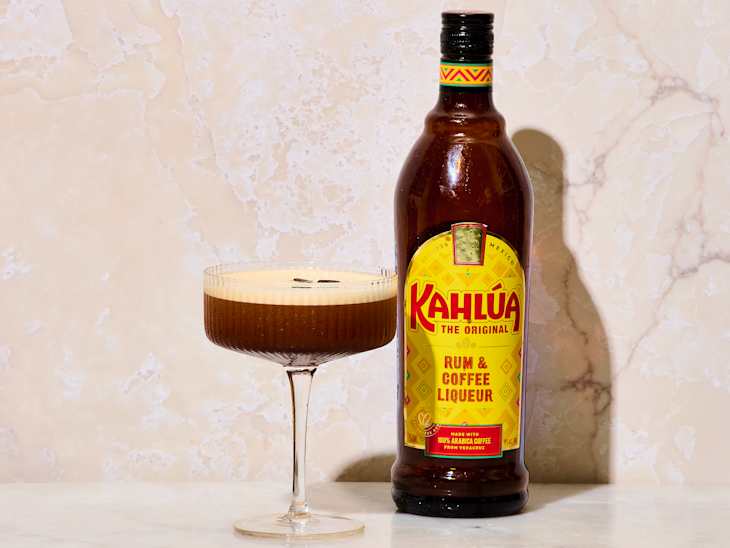 A glass of coffee cocktail topped with foam and coffee beans next to a bottle of Kahlúa liqueur on a marble surface.