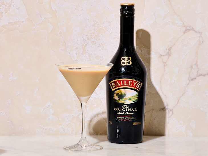 A martini glass filled with creamy beige liquid, garnished with coffee beans, next to a bottle of Baileys Irish Cream.