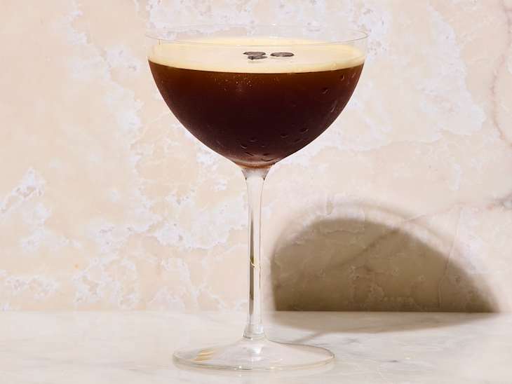 Cocktail in a coupe glass with a dark brown liquid topped with foam and coffee beans, set against a light marble background.