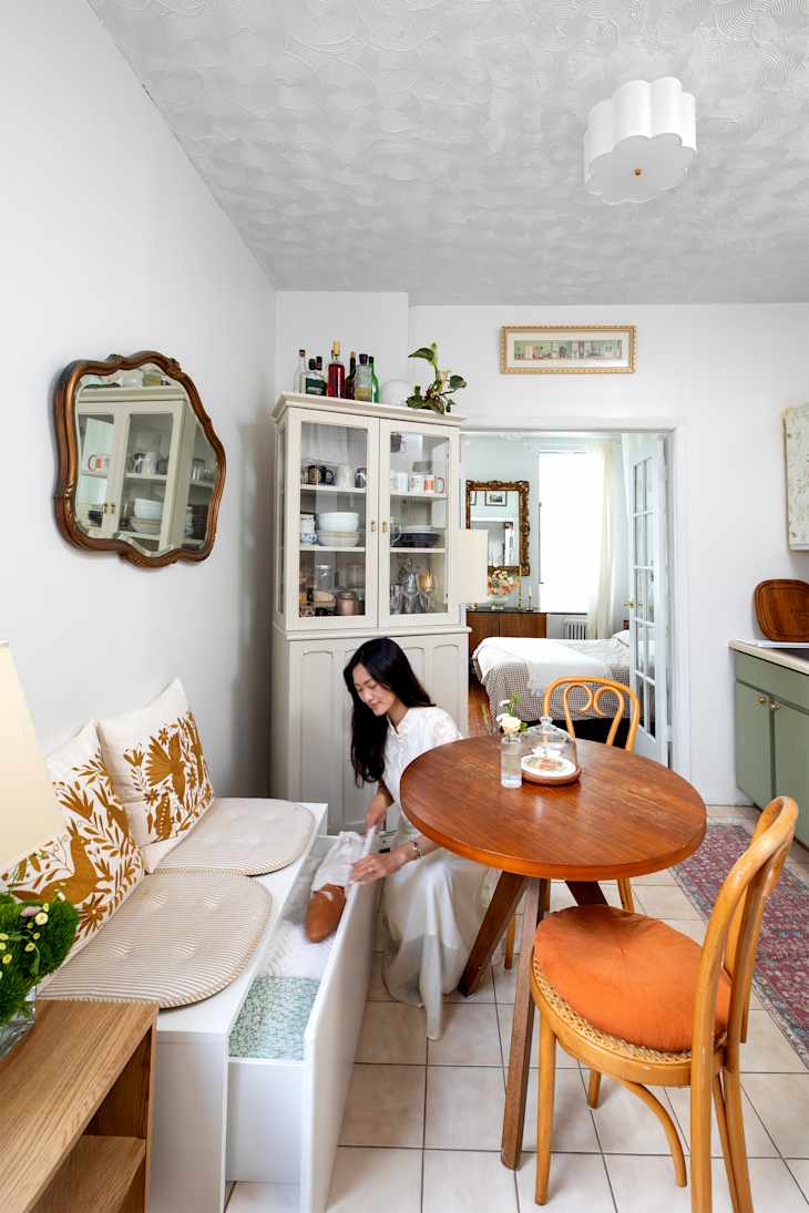 A dweller in a white dress organizes a storage bench in a cozy kitchen with a round wooden table, vintage decor, and a glass cabinet.