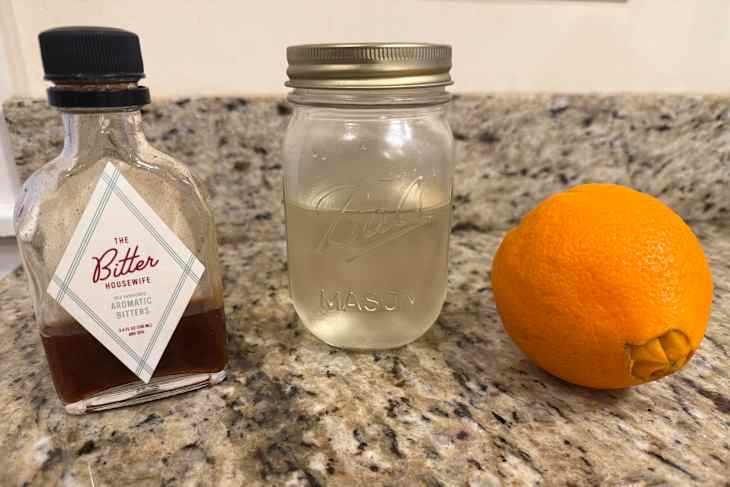 A bottle of aromatic bitters, a mason jar with liquid, and an orange on a granite countertop.