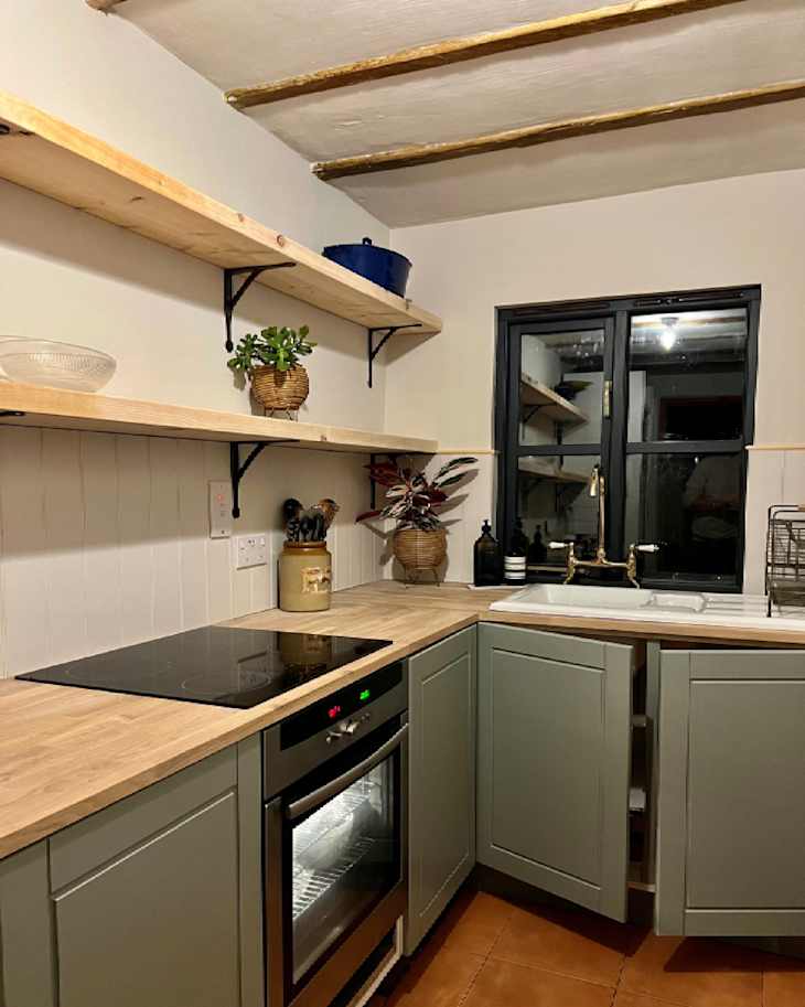 Modern kitchen with green cabinets, wooden shelves, a black cooktop, and a window above the sink.