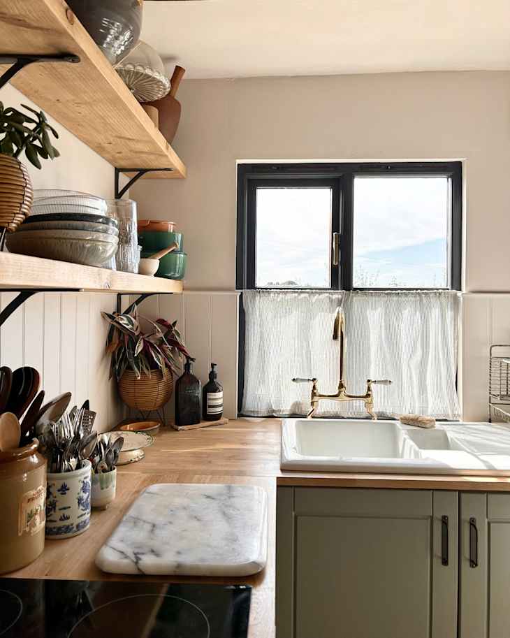 Bright kitchen with wooden shelves holding dishes, a marble cutting board, and a brass faucet over a white sink.