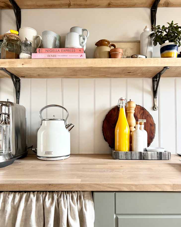 Bright kitchen countertop with a white kettle, wooden cutting board, yellow bottle, and various ceramic mugs and cookbooks on shelves.