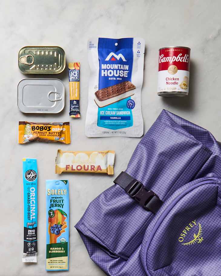 Assorted outdoor snacks including ice cream sandwich, beef stick, fruit jerky, and a purple Osprey backpack.