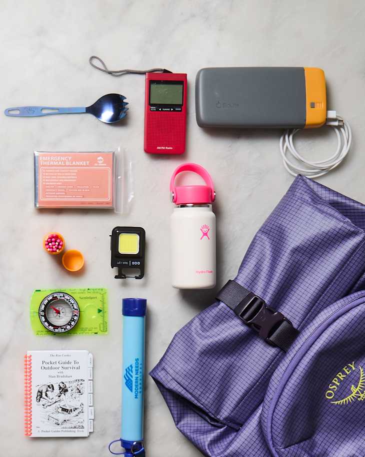Camping gear laid out on a surface, including a backpack, thermal blanket, radio, water bottle, compass, and survival guide.