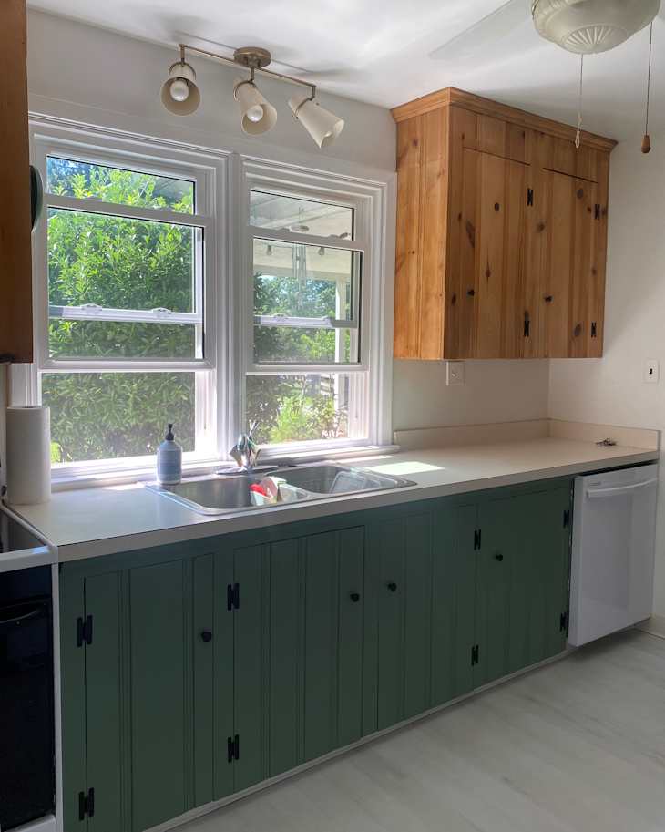 Bright kitchen with green cabinetry, double sink, and wooden upper cabinets, featuring natural light from two windows.