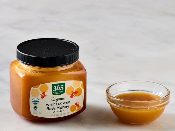Organic wildflower raw honey in a jar with a black lid next to a small glass bowl of honey.