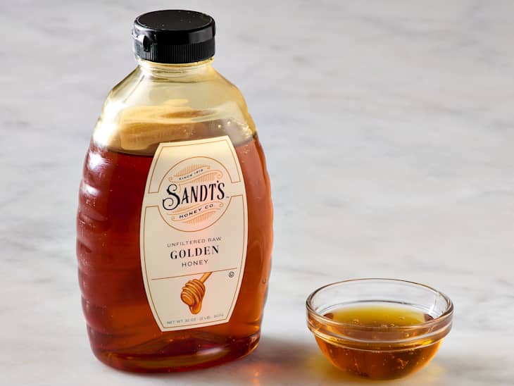 A bottle of unfiltered raw golden honey next to a small glass bowl of honey on a marble surface.