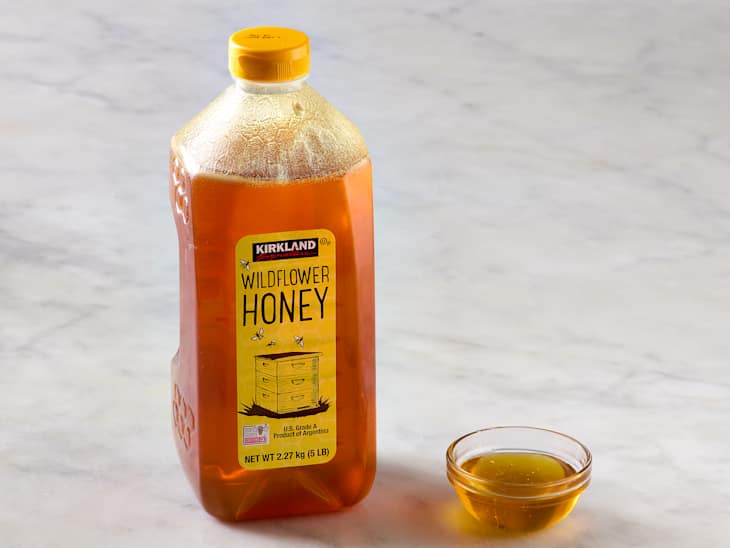 A bottle of wildflower honey with a yellow cap next to a small glass dish of honey on a marble surface.