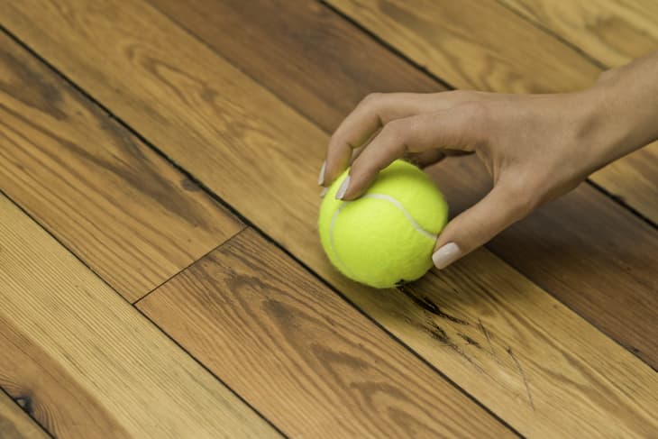 Hand holding a yellow tennis ball on a wooden floor.