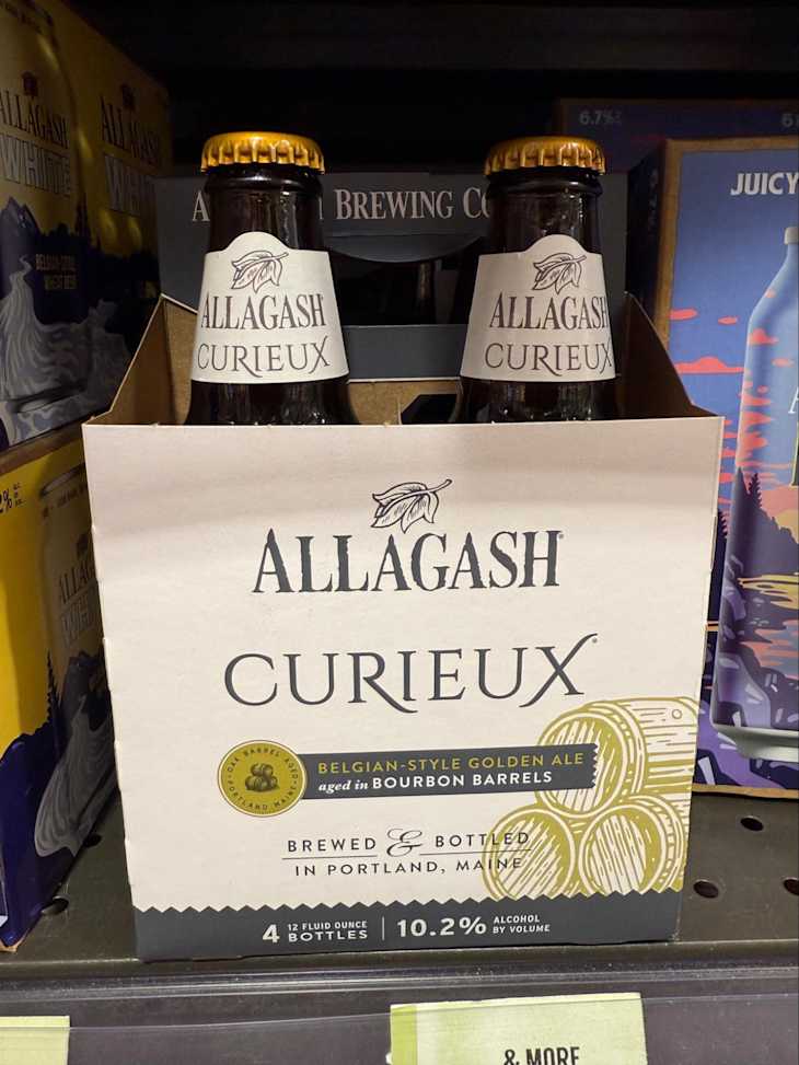 Four-pack of Allagash Curieux Belgian-style golden ale in bourbon barrel-aged packaging, displayed on a store shelf.