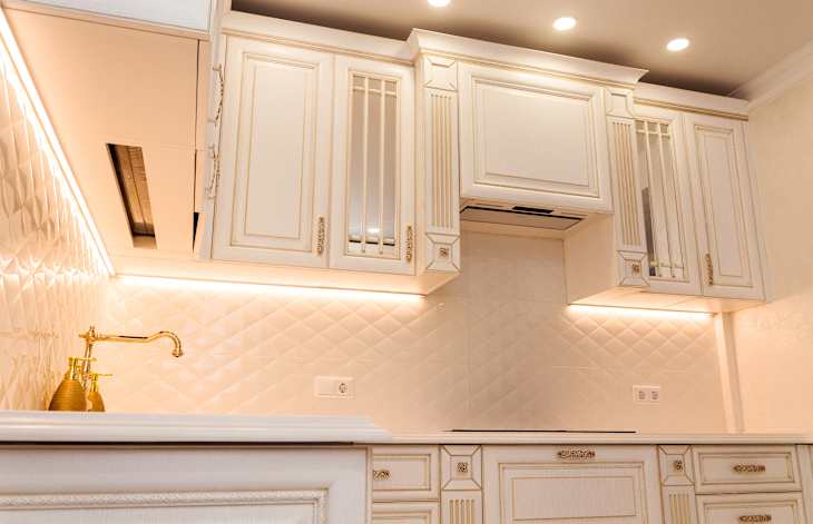 Luxurious white kitchen with gold-accented cabinets and modern lighting. Features tiled backsplash, integrated appliances, and under-cabinet lighting.