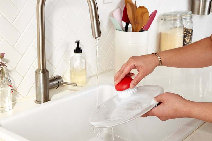 hand holding and scrubbing dish under water at kitchen sink using a red scrub brush
