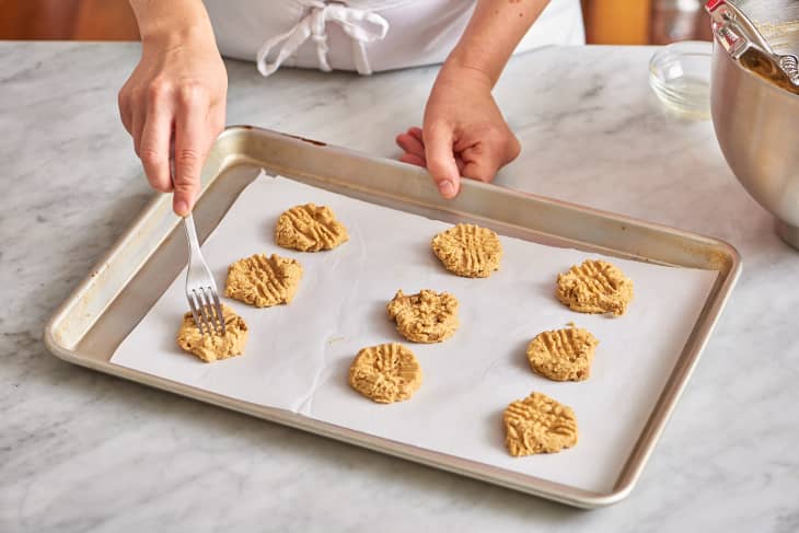 Person pressing fork marks into peanut butter cookie dough on a baking sheet.