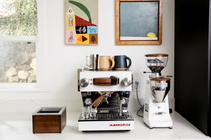 Espresso machine with mugs on top, next to a coffee grinder on a white countertop, with colorful wall art above.