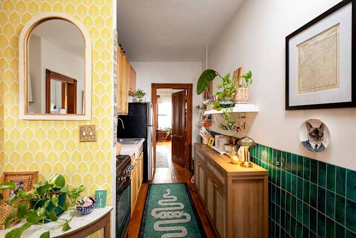 Narrow kitchen hallway with yellow patterned wallpaper, wooden cabinets, green tile backsplash, and decorative plants.