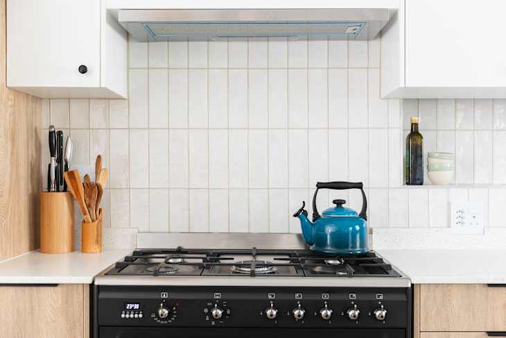 Bright blue kettle on a gas stove, black free standing oven, vertical textured wall tile, white top cabinets, pull out extractor fan, styled white counter tops