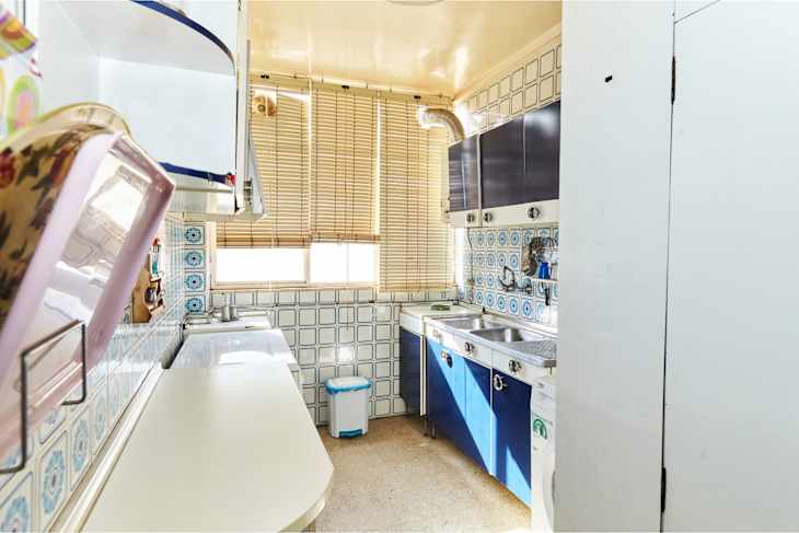 Compact kitchen with blue cabinets, tiled backsplash, sink, and window with blinds.