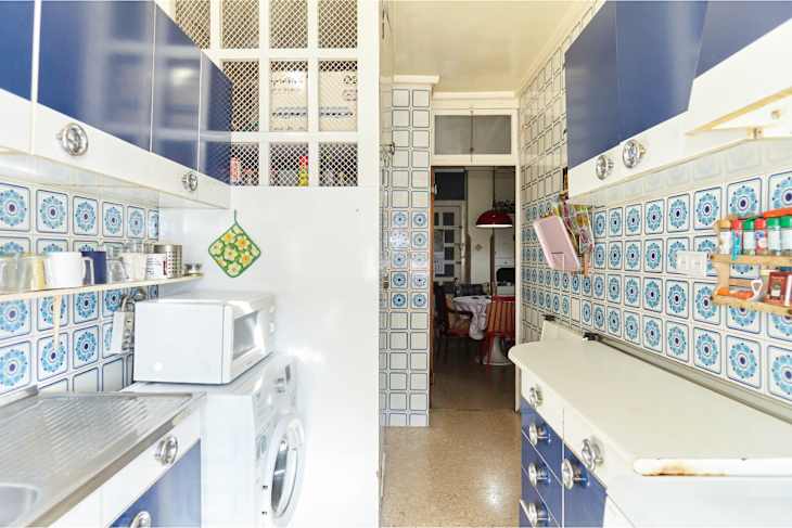 Narrow kitchen with blue cabinets, patterned tiles, a microwave, and a washing machine, leading to a dining area.