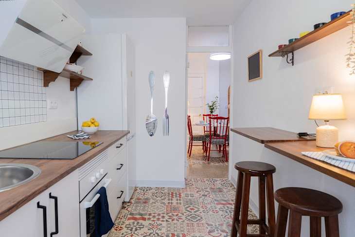 Modern kitchen with wooden countertops, white cabinets, decorative utensils on the wall, and a dining area visible in the background.