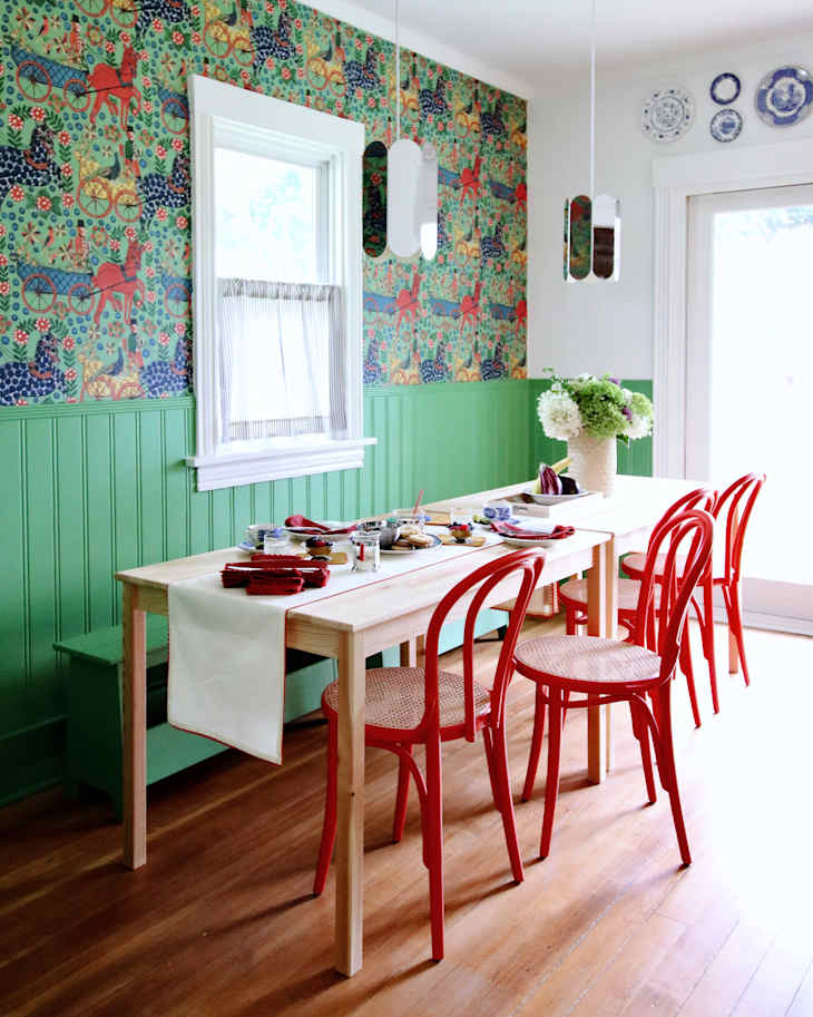 Bright dining room with a wooden table, red chairs, green wainscoting, floral wallpaper, and a vase of flowers.