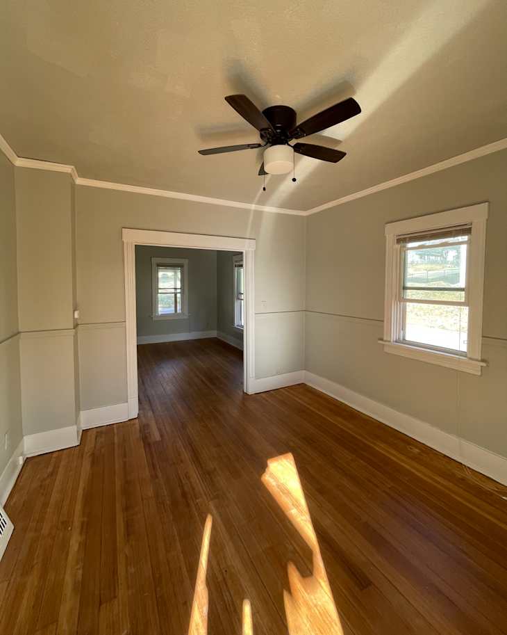Empty room with light green walls, hardwood flooring, a ceiling fan, and windows allowing natural light.