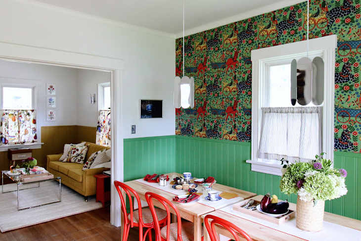 Bright dining area with green wainscoting, red chairs, floral wallpaper, and a table set with dishes and flowers.