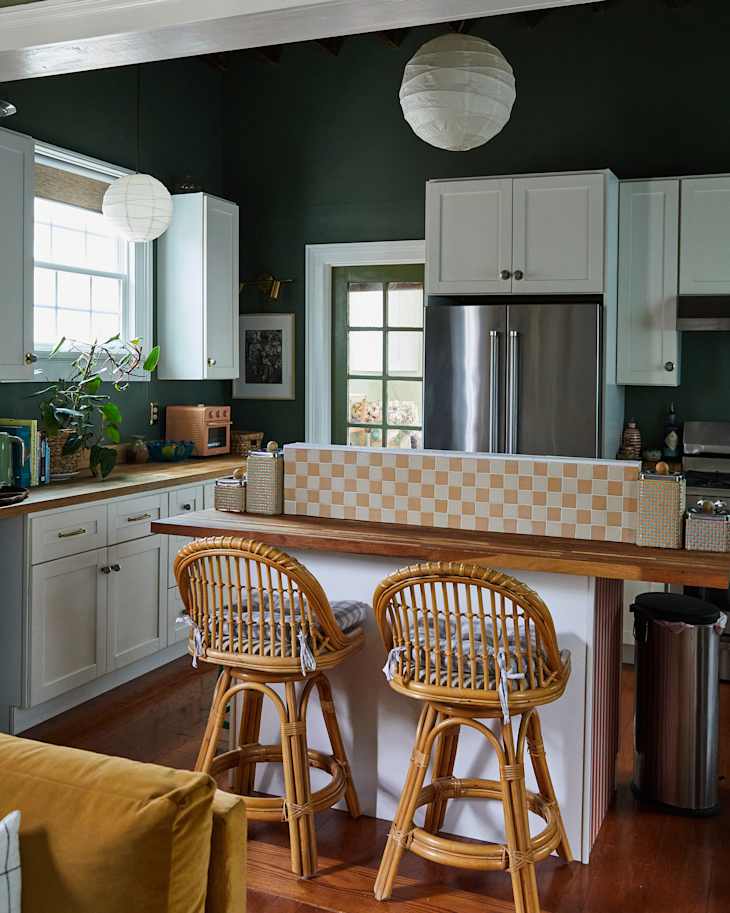 Cozy kitchen with dark green walls, wooden countertops, rattan bar stools, and a checkered backsplash.
