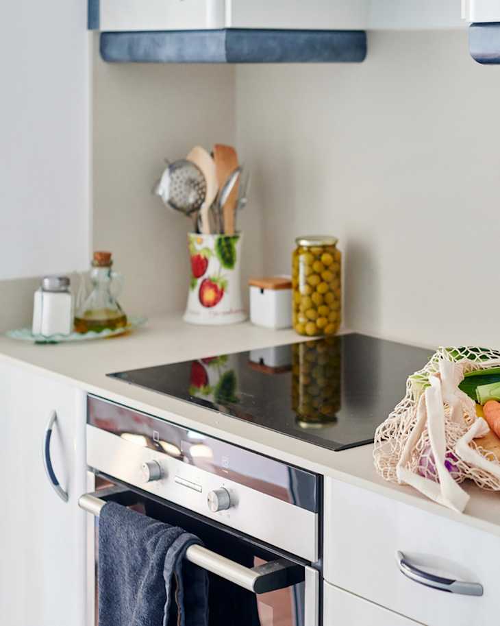 Modern kitchen with electric stovetop, utensils in a fruit-patterned holder, jar of olives, and vegetables in a mesh bag.