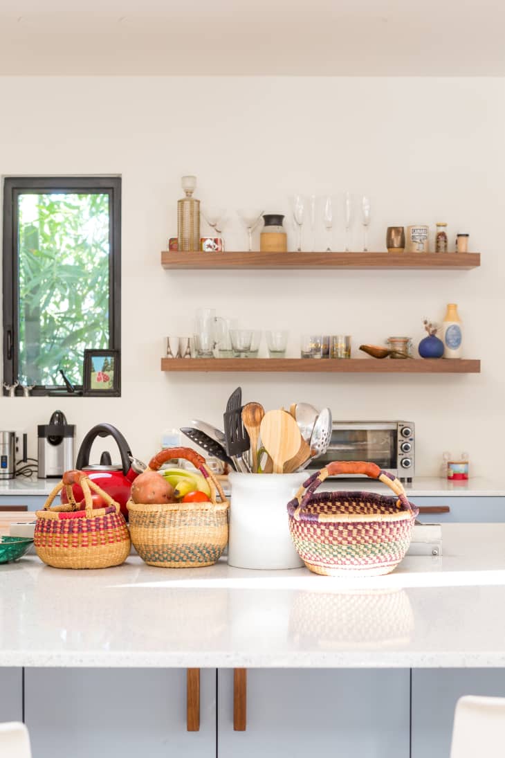 Kitchen with woven baskets of fruit, utensils in a white holder, and wooden shelves with glassware and decor.