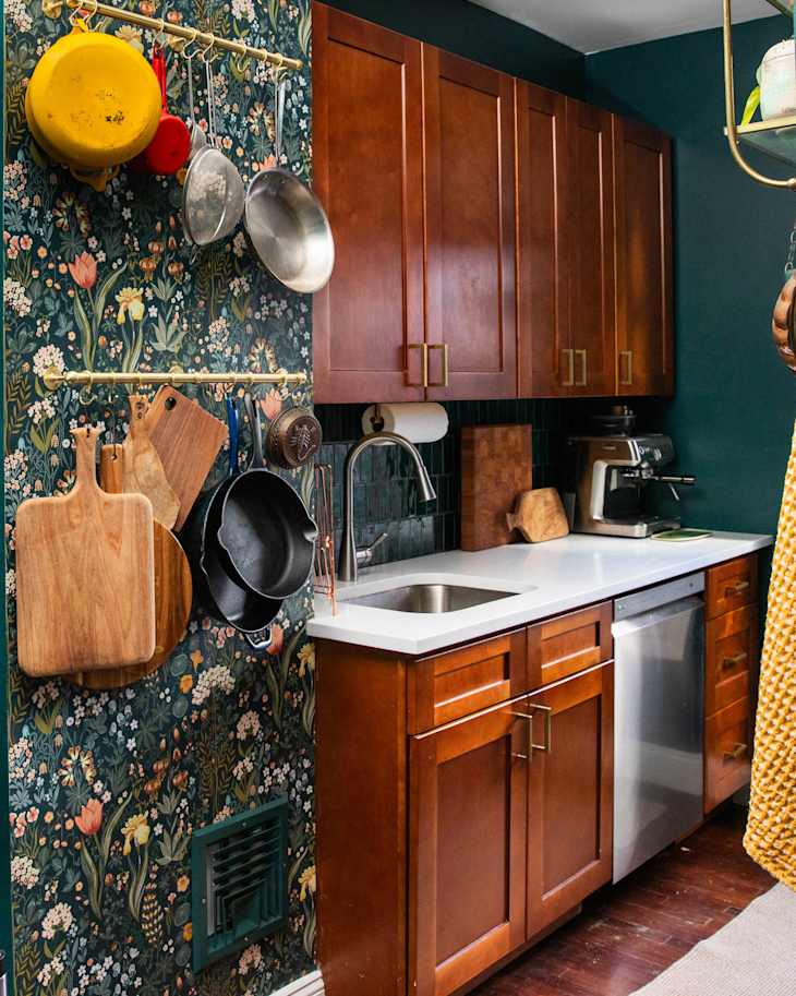 Cozy kitchen with dark green floral wallpaper, wooden cabinets, stainless steel appliances, and hanging cookware.