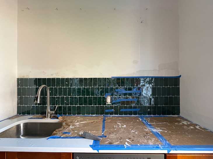 Kitchen countertop with a stainless steel sink, green tiled backsplash, and blue painter's tape for renovation.