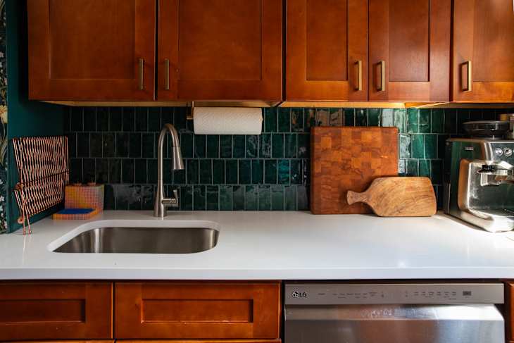 Modern kitchen with wooden cabinets, green tile backsplash, stainless steel sink, and cutting boards on a white countertop.