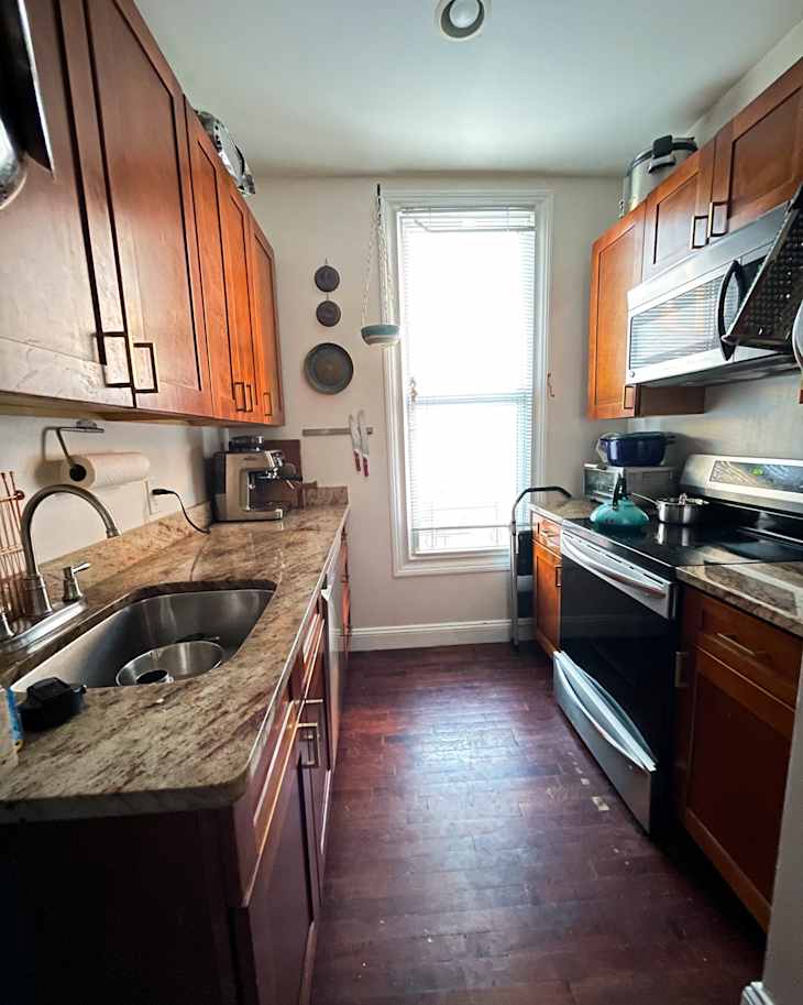 Narrow kitchen with wooden cabinets, granite countertops, stainless steel appliances, and a window with blinds.